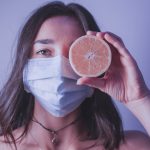 Young female in medical mask looking at camera and covering eye with half of orange while standing near wall during pandemic